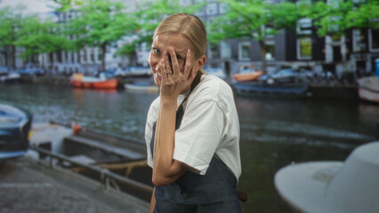 Woman in apron gently covers face with hand beside moored canal boats on a cobbled street, smiling through fingers; modesty playful warmth.