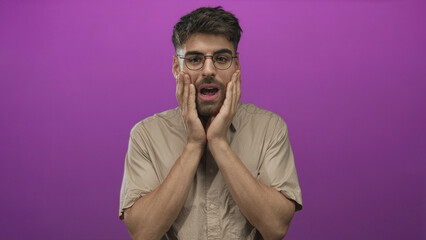 Man wearing glasses and beige shirt presses both hands to cheeks with open mouth and raised eyebrows in purple studio; surprise.