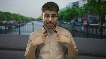Young hispanic man wearing beige shirt pointing finger to chest on street by canal; pride.