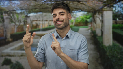 Casual bearded smiling man pointing index finger toward ornate stone building in green park; optimism.