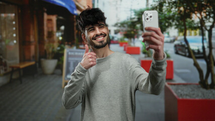 Young man taking selfie with smartphone on restaurant terrace with plants in urban street showing thumbs up and smiling naturally in an outdoor setting