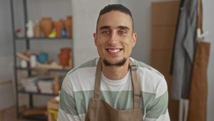 Man potter in apron smiling and showing teeth in a pottery studio with clay pots and shelves behind him; craft pride.