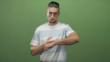 Man points finger to wrist and taps bare wrist in studio with green backdrop, wearing striped tshirt and visible ring on left hand; impatience time reminder.