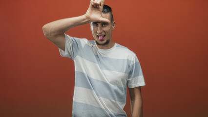 Man forms l with hand on forehead and sticks tongue out in a studio with orange backdrop wearing a...