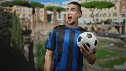Man holding soccer ball with right hand, mouth open, wearing blue and black striped jersey, standing near ancient building ruins in city; excitement celebration.