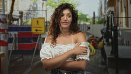 Woman with arms crossed holding shoulder in white off shoulder blouse on street beside construction barriers and warning sign; confidence resilience.