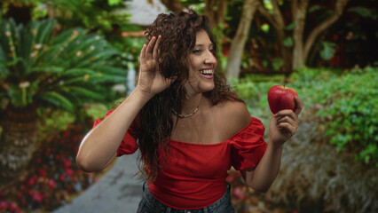 Obraz premium Woman holding red apple with hand cupped to ear, bare shoulder visible on a park path among greenery and flowers; playful curiosity.