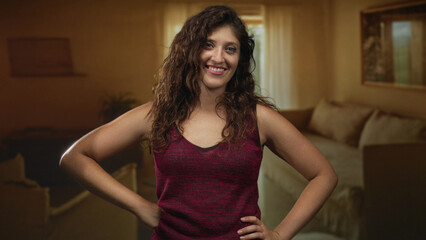 Fototapeta premium Woman with hands on hips and bare arms visible in a living room inside a building, smiling at camera; confidence comfort.