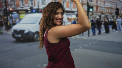 Naklejka premium Young woman flexes bare upper arm and smiles while posing amid a busy city street with pedestrians, cars and shopfronts visible; confidence.