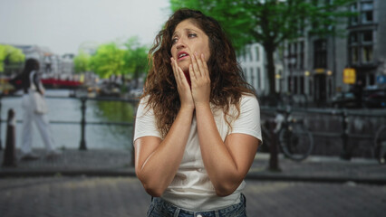 Fototapeta premium Hispanic woman with hands on cheeks, freckled face and white t shirt standing on street by canal in amsterdam; urban anxiety.