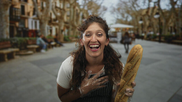 Woman holding a baguette and wearing apron and plastic gloves on a bustling street park with benches and trees; local bakery joy. - Powered by Adobe