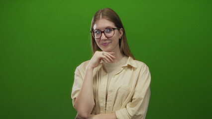 Young blonde woman wearing glasses touches chin with subtle smile in studio with green backdrop...