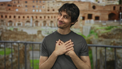 Fototapeta premium Man in casual grey shirt touches chest and looks at camera amid ancient roman ruins at building; sincerity.