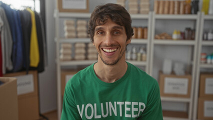 Man wearing green volunteer t shirt smiles beside stacked food donation boxes on metal shelves in storage room building; compassion.