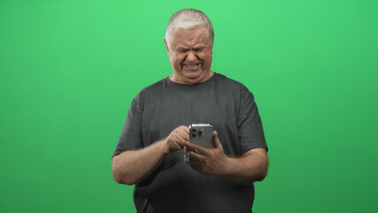 Man tapping smartphone with right hand, holding phone in left hand and squinting at screen in green screen studio; frustration.