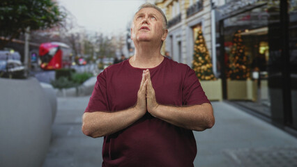 Fototapeta premium Senior man with hands pressed in prayer at chest on a busy city street sidewalk beside a storefront and christmas tree, looking upward in maroon shirt; faith hope.