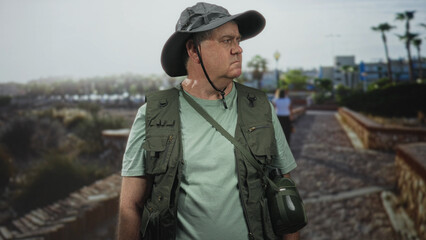 Man in a fishing vest holding a canteen and looking to the right on a seaside promenade; serenity solitude.