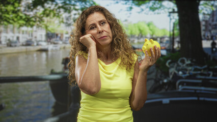 Middle age hispanic woman in yellow top holds a yellow piggy bank and rests cheek on fist by a canal on a street with bicycles and water; financial worry.