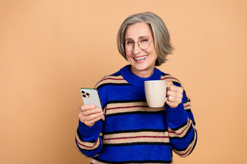 Happy mature woman with glasses in a blue striped sweater holds a mug and checks her smartphone against a warm beige background