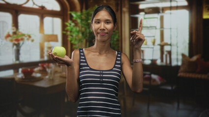 Thai woman holding a green apple in her palm and pointing one finger up while smiling inside a...