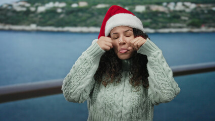Woman wearing santa hat by the beach with a sad expression, dressed in warm sweater, showcasing emotions with the sea in the background on a cloudy day. © Krakenimages.com