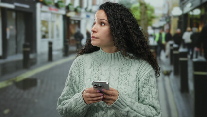 Woman in sweater using smartphone on rainy street, focused and thoughtful, surrounded by blurred pedestrians and urban elements, representing modern communication outdoors.