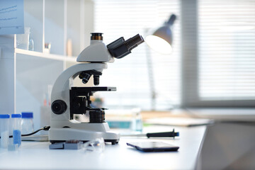 Laboratory microscope standing on desk surrounded by scientific equipment and glassware, sunlight...