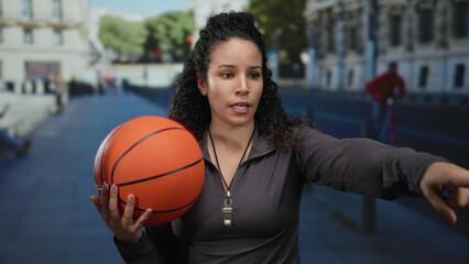 Woman holding basketball standing on urban street pointing outdoors in daytime wearing gray jacket with curly hair captured in lively hispanic environment.