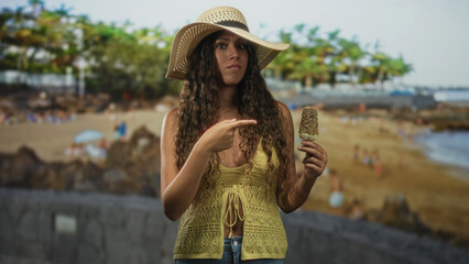 Woman pointing finger at a popsicle while holding it near her chest on a street promenade by a sandy beach with palm trees; curiosity.