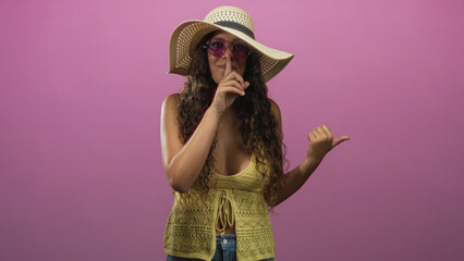Hispanic woman in straw sunhat and purple sunglasses points thumb to side then makes finger to lips shush in studio; playful secrecy.