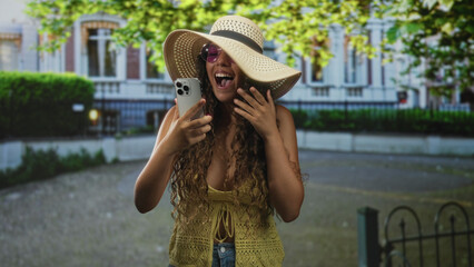 Fototapeta premium Hispanic teenage woman wearing sunhat and sunglasses holds smartphone, laughs with mouth open and covers face with hand in street; joy connection.
