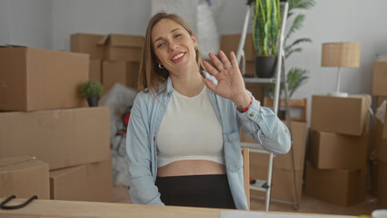 Woman with pregnant belly waves at camera in new apartment interior; anticipation joy.