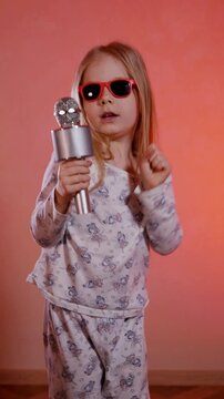 Vertical. Aspiring pop star in patterned pajamas belts out a tune and dances funny. Young girl in red sunglasses sings passionately into a silver microphone against a soft peach background.