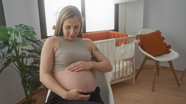 Pregnant woman sitting in a cozy baby room, smiling and gently holding her belly, surrounded by nursery decor, emphasizing a serene indoor atmosphere at home. - Powered by Adobe