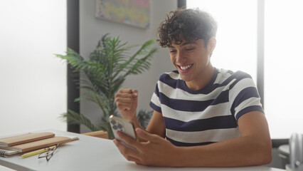 Teenage boy holds smartphone and makes a fist pump at a study desk inside a building  joy achievement success. © Krakenimages.com
