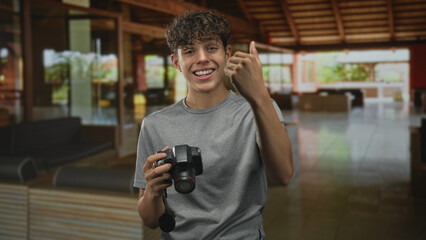 Man holding dslr camera making pointing and thumbs up gestures in resort building lobby while smiling and moving his hand; playful confidence.