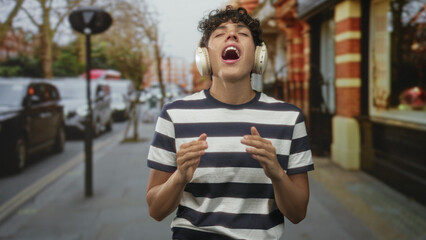 Teenage boy in striped t shirt with over ear headphones singing and holding hand to chest while walking on a street lined with parked cars and brick shops; joy.