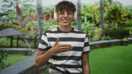 Fototapeta premium Smiling teenage boy wearing striped t shirt and over ear headphones, hand palm up to chest gesture in forest setting with palm trees and stone wall; playful joy.