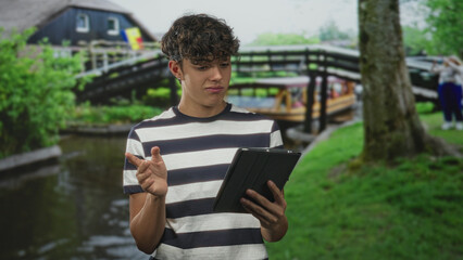 Teen man holds tablet with one hand and presses palm to chest while reading a startling notification on a canal street with wooden bridge nearby; surprise reflection.