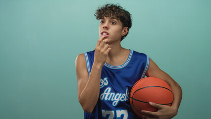 Teenage boy holding basketball, finger to lips for silence, wearing blue los angeles jersey in studio; thoughtful reflection quiet focus.