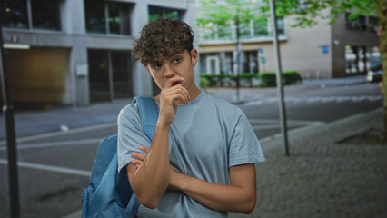 Fototapeta premium Teen boy with backpack places finger to lips on urban street by buildings, sidewalk, lamppost and storefront, arms crossed and looking pensive; thoughtful curiosity.