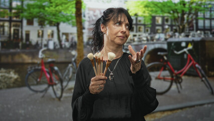 Woman holds makeup brushes to chest, hands clasped, wearing black dress on street; quiet reflection.