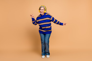 Fototapeta premium Happy mature woman wearing striped knitwear dancing with yellow headphones in a beige studio background