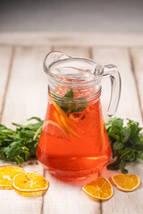 Red citrus lemonade in a glass pitcher with mint, orange slices and fresh herbs on rustic wooden background