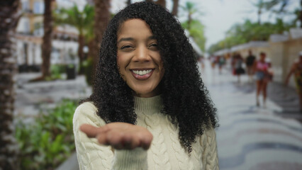 Woman smiles and blows kiss with hand extended on sunny city street lined with palm trees  affection warmth connection. © Krakenimages.com