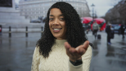 Woman smiling with curly hair extends her hand forward on a wet street flanked by historic...