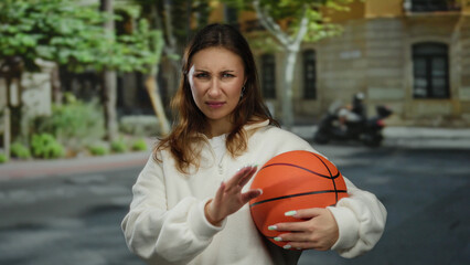 Young caucasian woman holding basketball making rejection gesture on city street outdoors,...