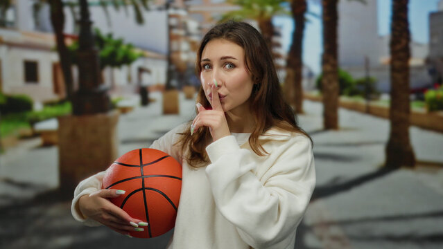 Young woman holds basketball gesturing silence outdoors in a park with palm trees and buildings in the background under a bright sky - Powered by Adobe