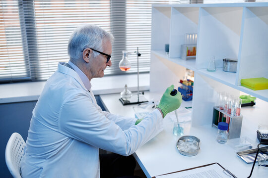 Senior Caucasian man working in laboratory, wearing lab coat and gloves, using pipette to transfer liquid into beaker, surrounded by scientific glassware and equipment on desk