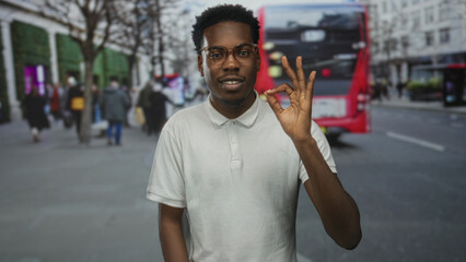 Young african american man in white t shirt shows ok gesture with hand on busy city street; approval.
